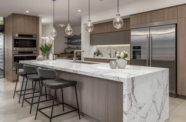 Luxury contemporary kitchen featuring a white marble waterfall island, natural wood cabinetry, integrated appliances, stainless steel fridge and elegant glass pendant lighting.