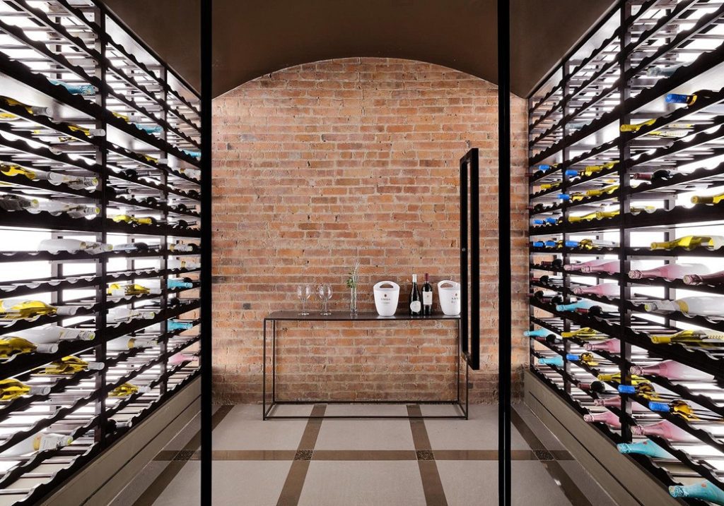 Wine cellar with black metal racks holding numerous wine bottles against a brick wall