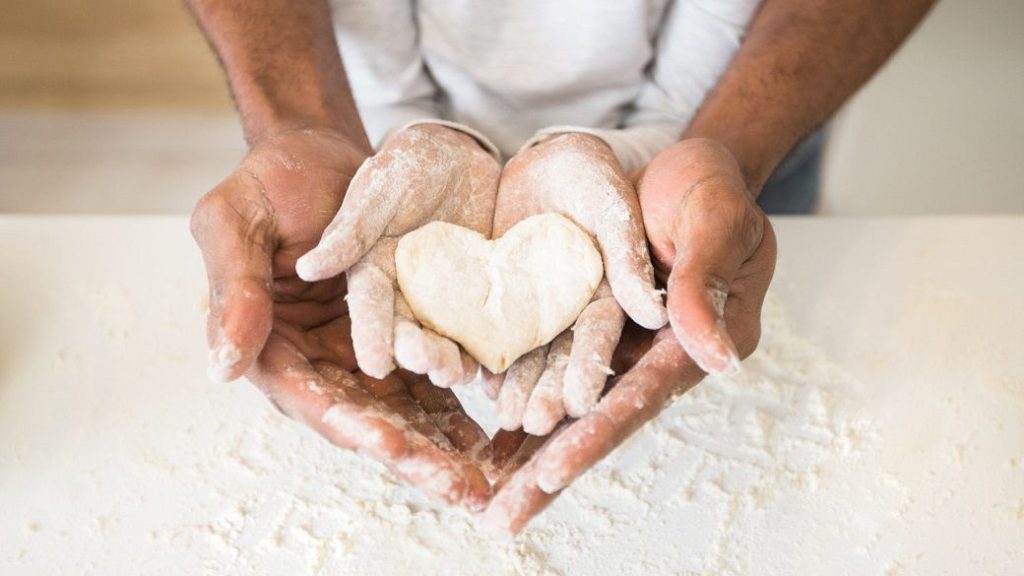 Two pairs of hands holding heart-shaped dough over a floured surface