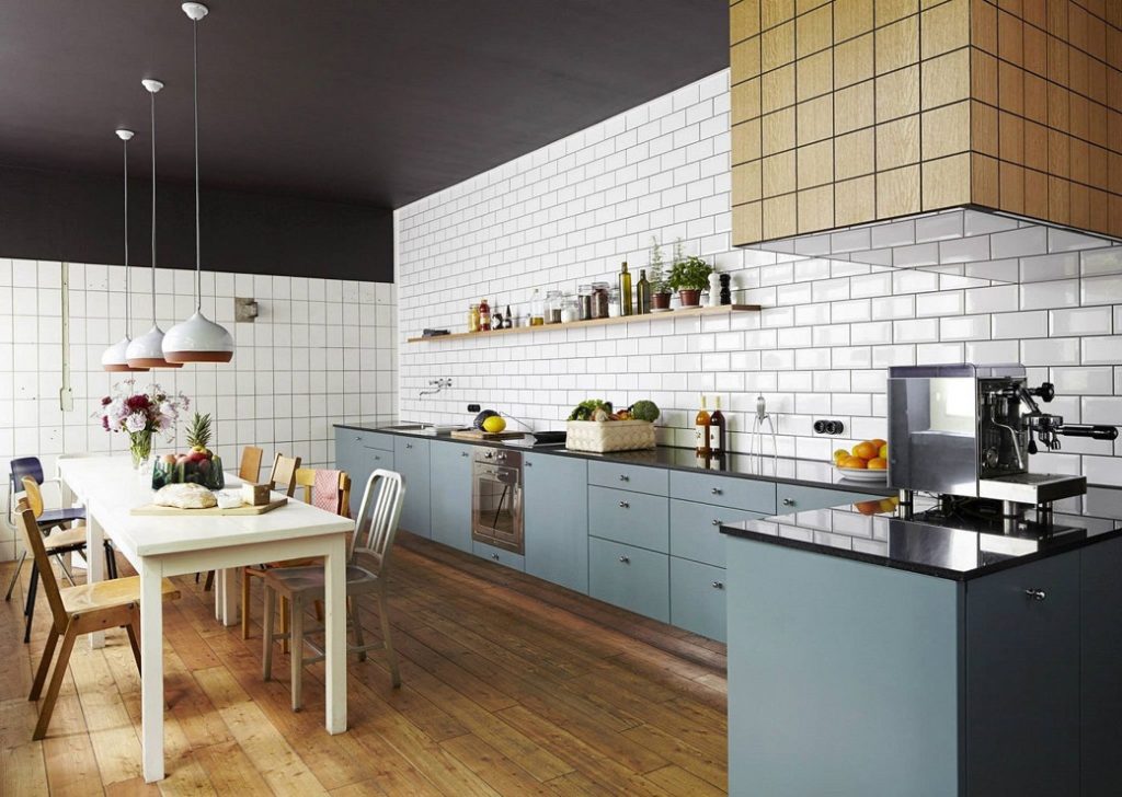 Long kitchen with blue cabinetry, white tiled walls, wooden floor, and dining table with mixed chairs