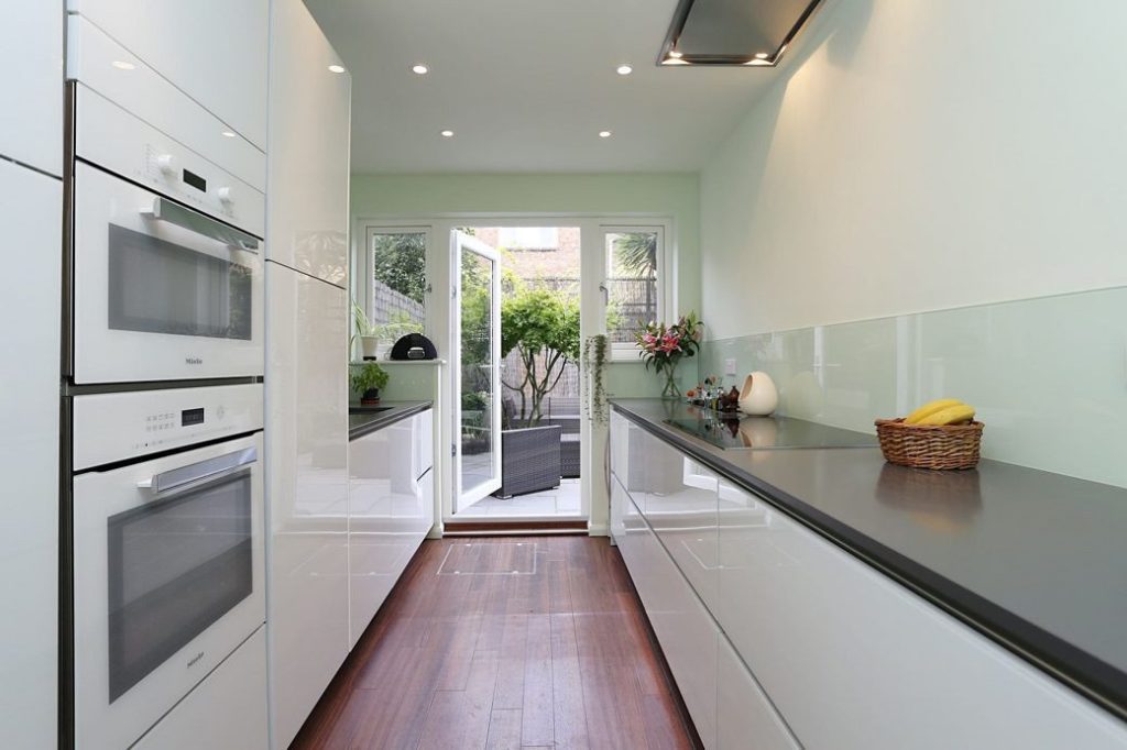 Bright galley kitchen with white cabinetry, dark countertops, wooden floor and glass door to garden
