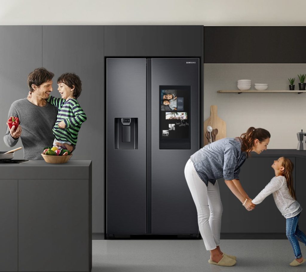 Family of four in a kitchen with dark cabinetry and a large black fridge with digital screen