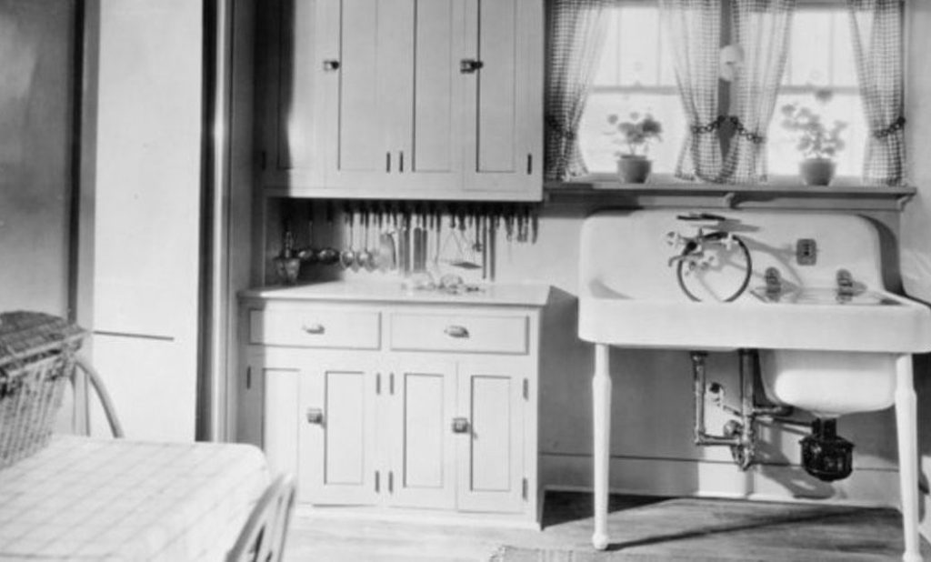 Black and white image of a kitchen corner with cupboards, a large sink, and a window with curtains