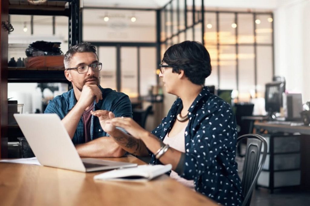 Two people discussing work at a table with a laptop and notebook in a modern office