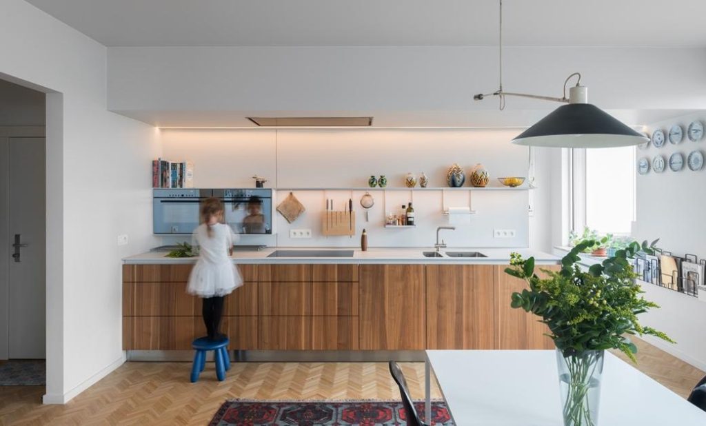 Child standing on blue stool at wooden kitchen cabinets with white countertop and built-in appliances