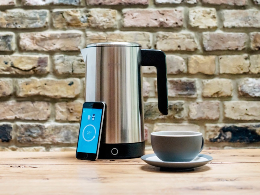 Stainless steel electric kettle next to a smartphone and a grey cup with saucer on a wooden table, brick wall background.