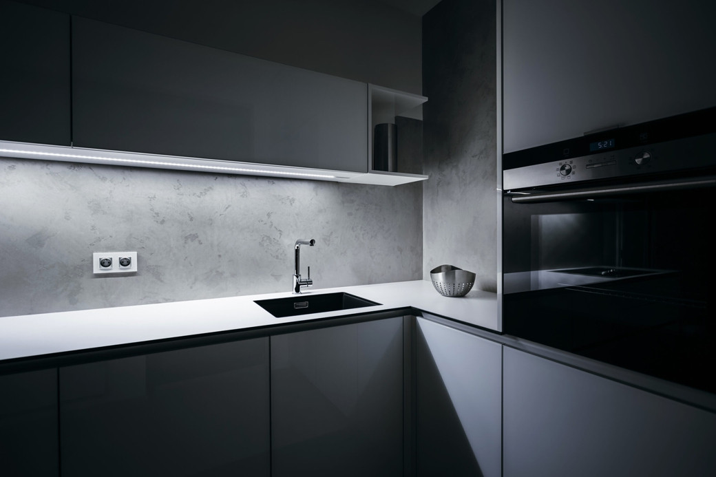 Modern minimalist kitchen corner with white countertops, gray cabinets, built-in oven, and under-cabinet lighting.
