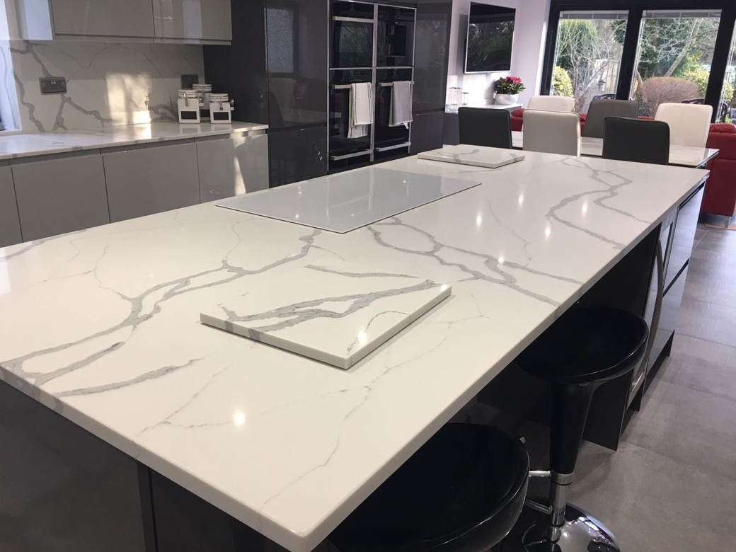Modern kitchen island with white marble-patterned countertop, black bar stools, grey cabinets, and built-in ovens in the back