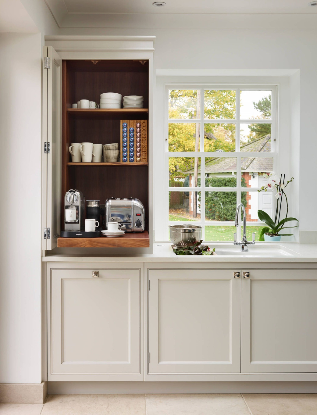 Open kitchen cupboard with coffee machine, toaster, mugs, plates, coffee pods, by window with a sink and orchid plant on coun