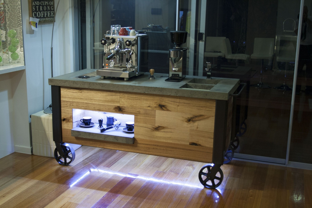 Wooden coffee station on wheels with a coffee machine, grinder, and illuminated shelf with cups and tools.
