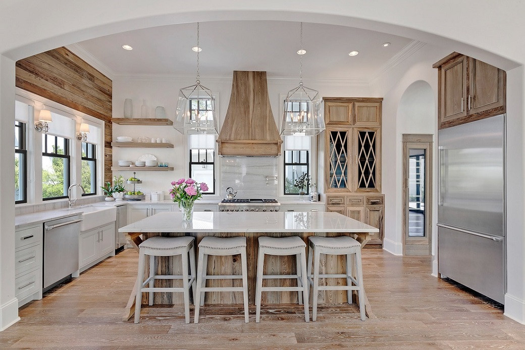 Bright farmhouse kitchen with wooden floors, central island with four stools, wood cabinetry, and large windows.