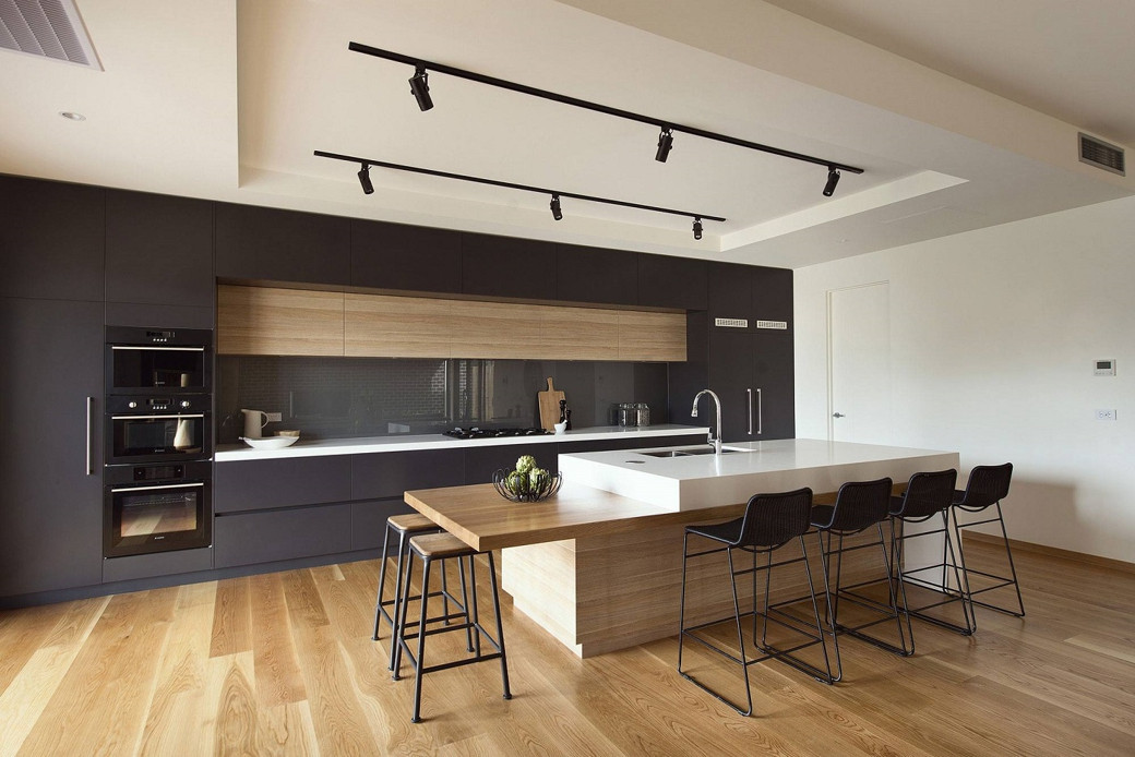 Modern kitchen with a wooden and white island, black stools, dark cabinetry, wooden floor, and track ceiling lights.