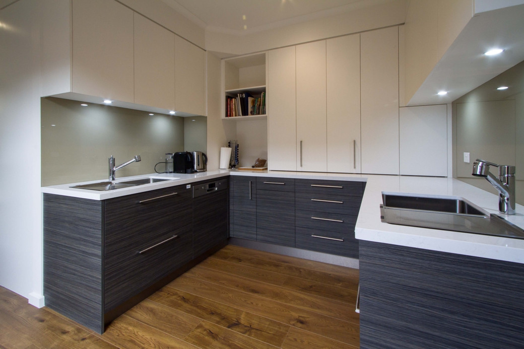 Modern kitchen with white upper cabinets, dark lower cabinets, wooden floor, two sinks, and under-cabinet lighting.