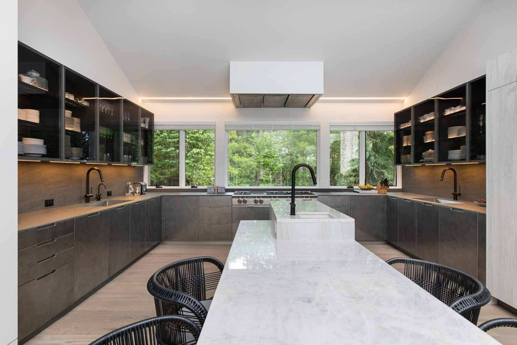Modern kosher kitchen with U-shaped dark grey cupboards, marble island, two black sinks, and large windows showing greenery o