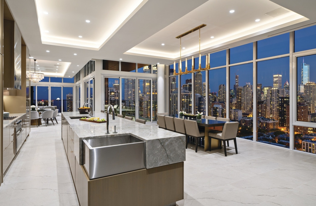 Spacious luxury penthouse kitchen with marble island, stainless steel sink, dining area, and city skyline view at dusk.