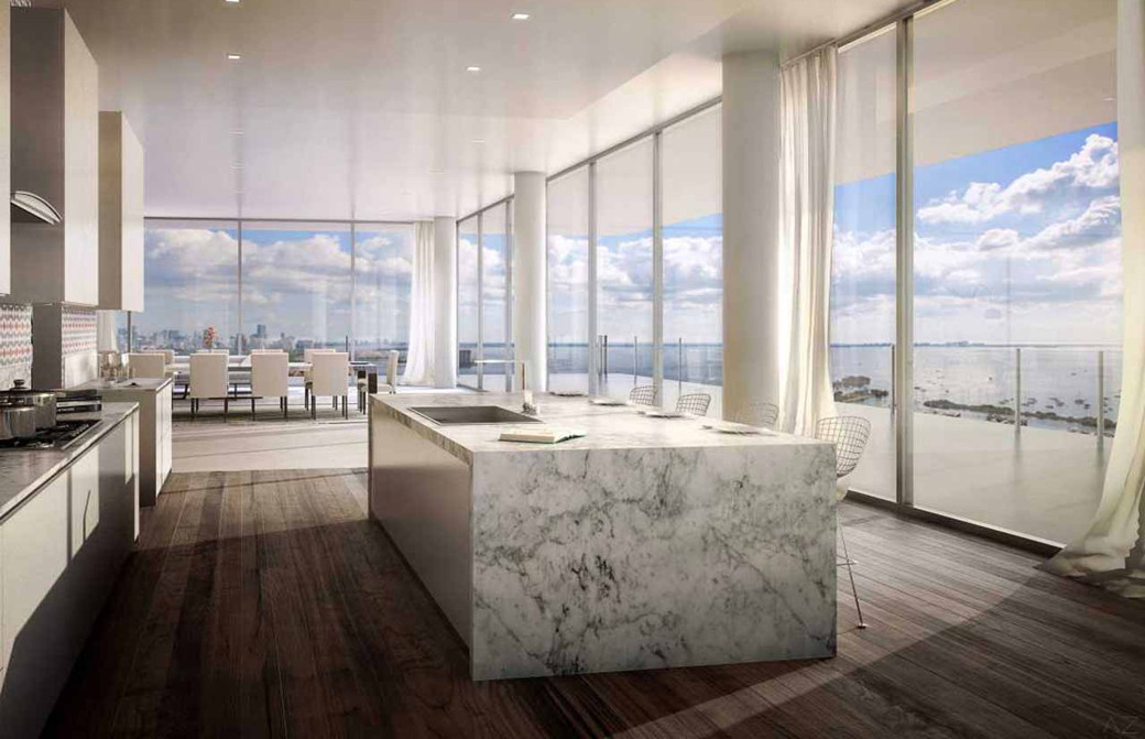 Open-plan penthouse kitchen with large white marble island, wood floors and floor-to-ceiling windows overlooking ocean.