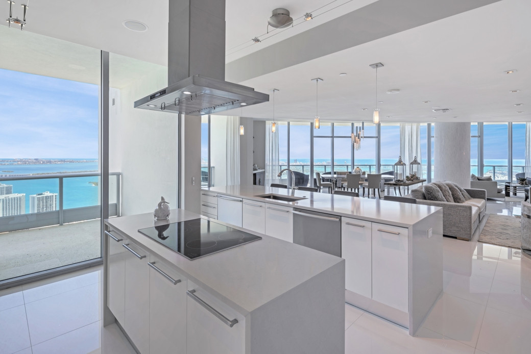 Bright modern penthouse kitchen with white countertops, island stove, and ocean views through floor-to-ceiling windows.