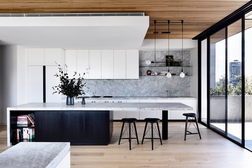 Modern penthouse kitchen with white and black cabinetry, marble countertops, wooden slatted ceiling, bar stools, and floor-to