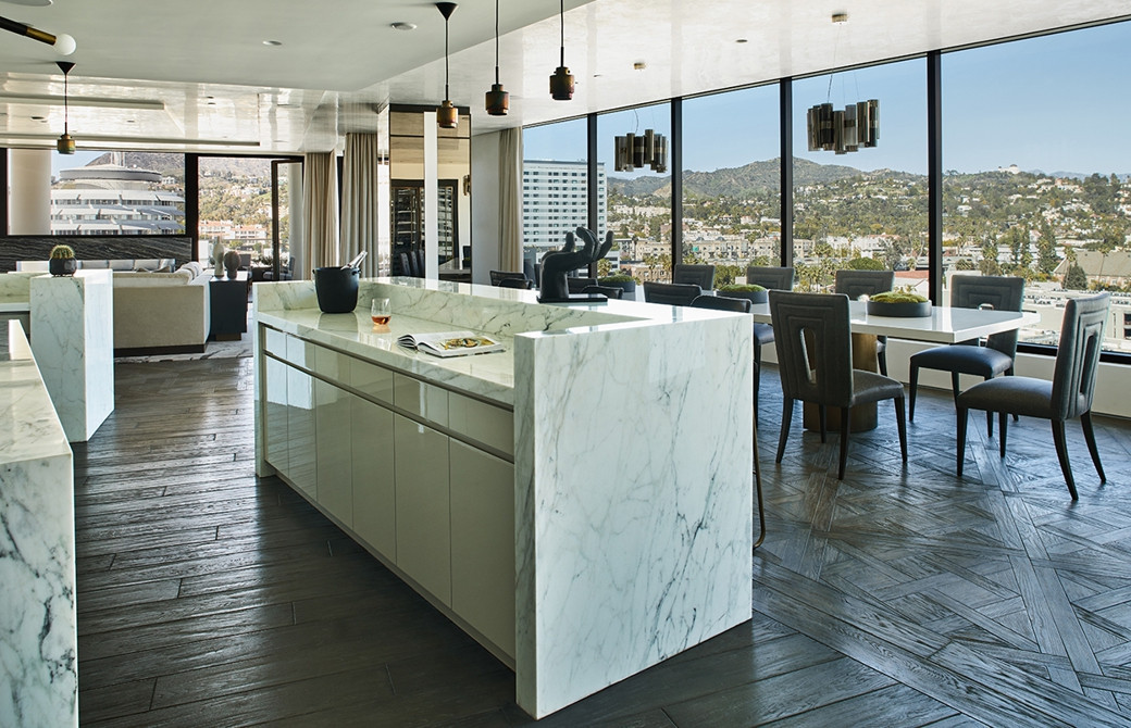 Open-plan penthouse kitchen with white marble island, dark wood flooring, grey dining chairs, and large floor-to-ceiling wind