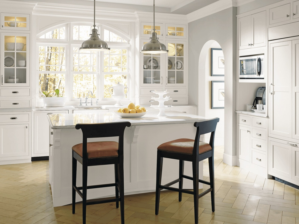 Bright white kitchen with marble island, two dark wood stools with brown seats, glass-front cabinets, and large windows.
