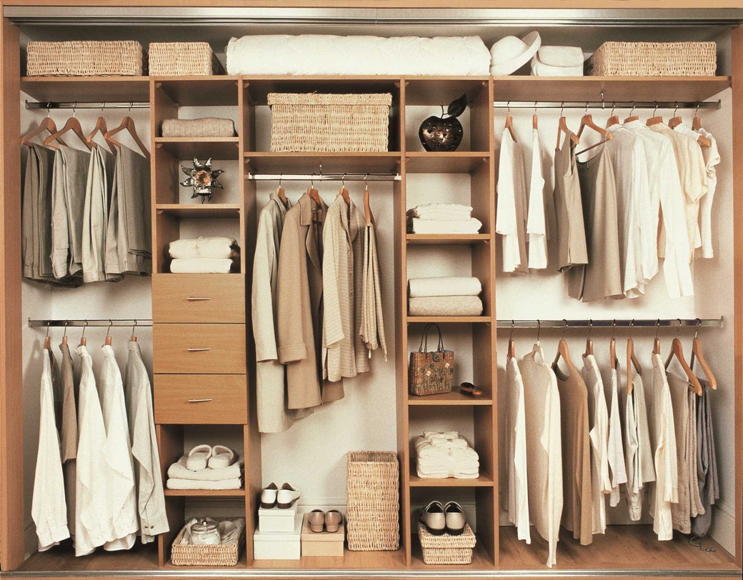 Organised wooden wardrobe with beige and white clothes on hangers, baskets, folded towels, shoes, and decorative items.