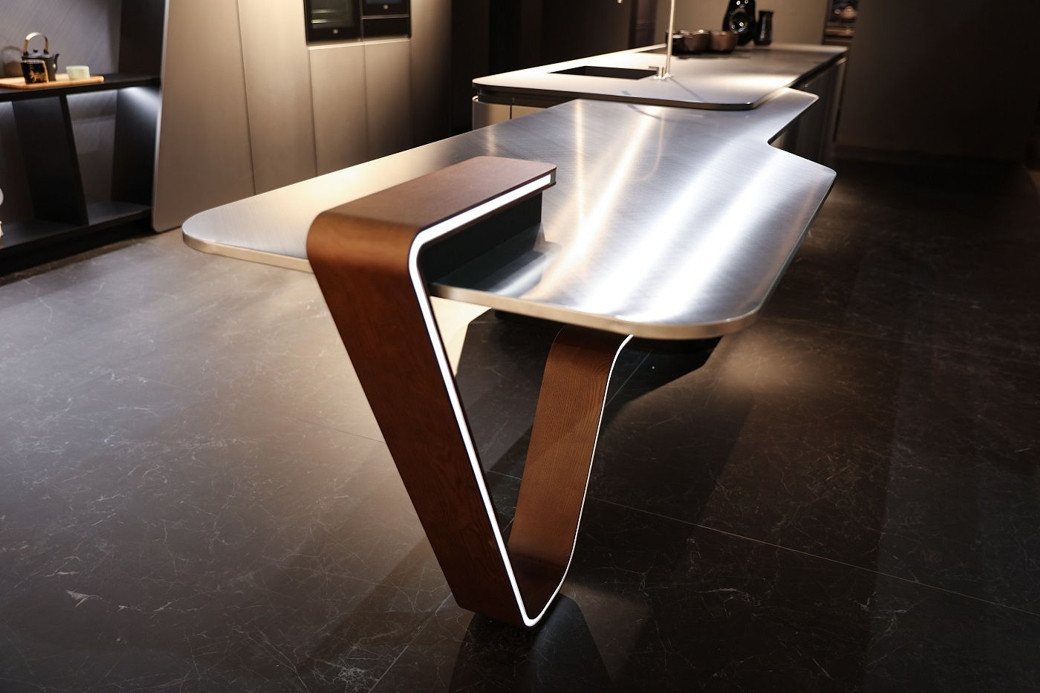 Ultra-modern kitchen island with a stainless steel top and sculptural wooden leg on dark tiled floor.
