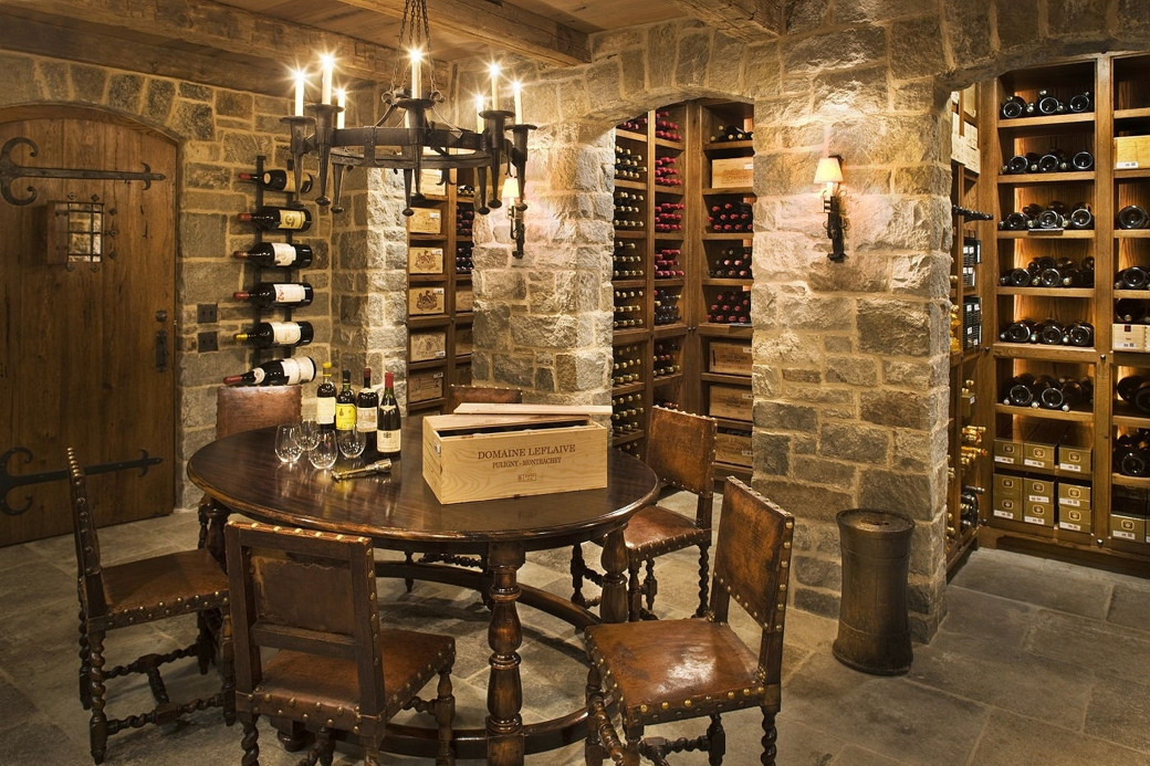 Rustic wine cellar with stone walls, wooden racks filled with wine bottles, round table with chairs, and chandelier.