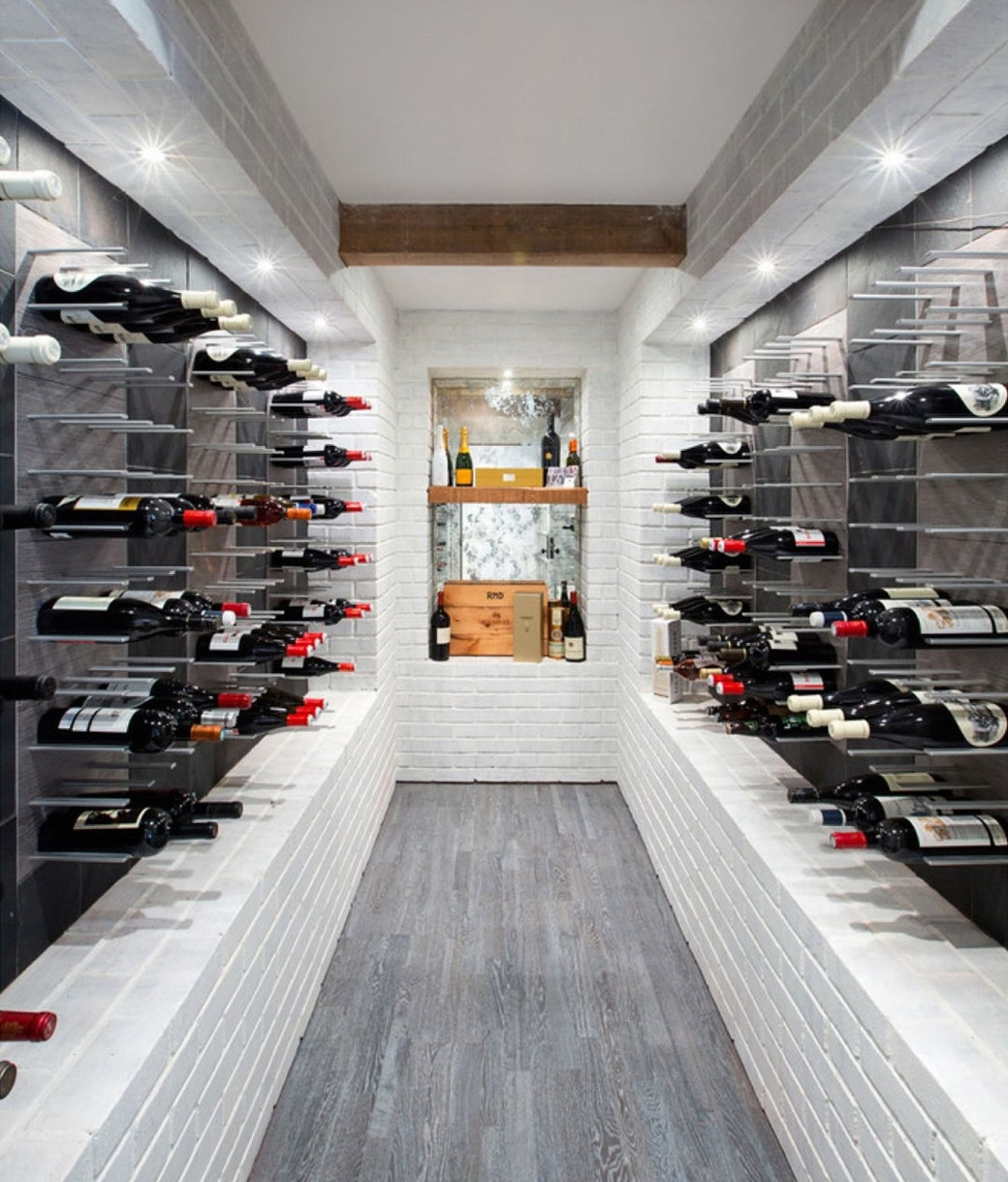 Modern wine cellar with white brick walls, metal racks holding wine bottles on both sides, grey wooden floor.