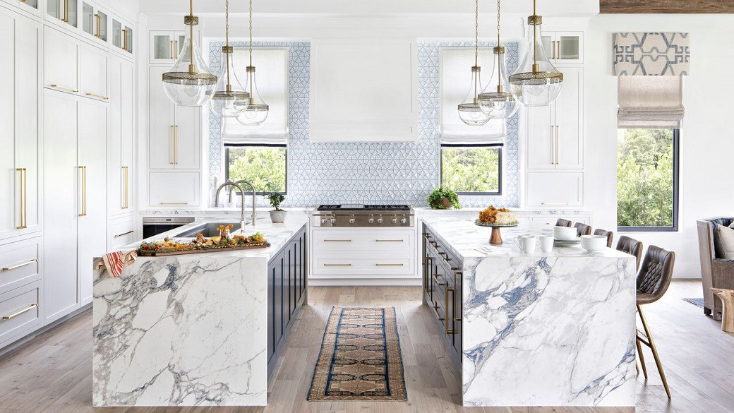 Modern kitchen with two rectangular marble islands, navy cabinets, brass handles, pendant lights, and patterned blue tile bac