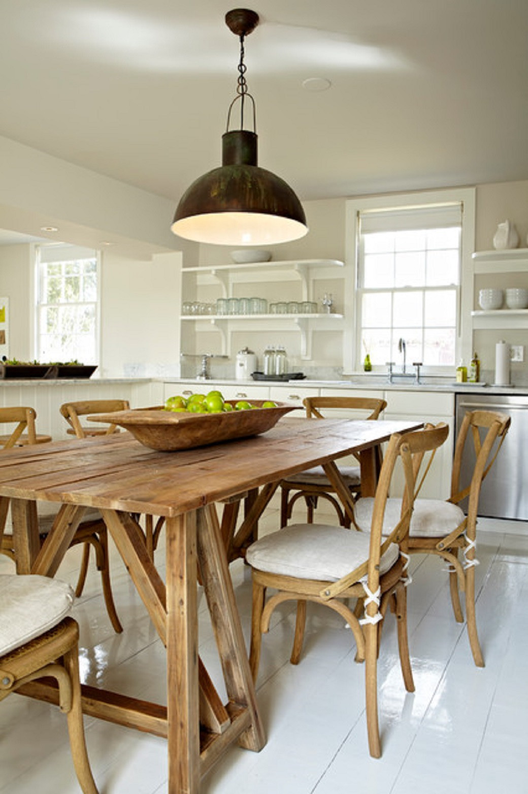Wooden dining table with six wooden chairs, a bowl of green apples, large hanging lamp, white kitchen with shelves and window