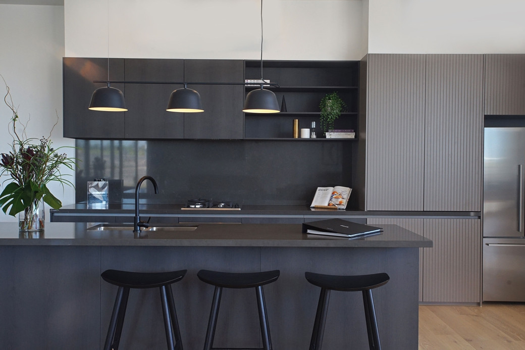 Modern kitchen with dark grey Caesarstone countertops, black stools, hanging pendant lights, and stainless steel fridge.