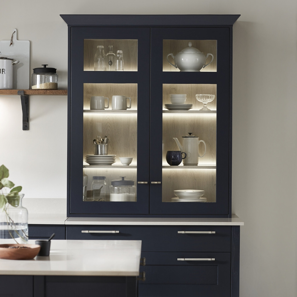 Kitchen cabinet with dark blue frames and glass doors showcasing dishes and glassware, lit with warm interior lighting.