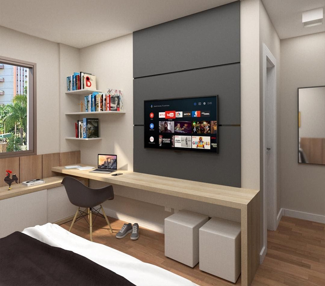 Small apartment bedroom with a built-in wooden desk, laptop, bookshelves, wall-mounted TV, and two white ottomans.