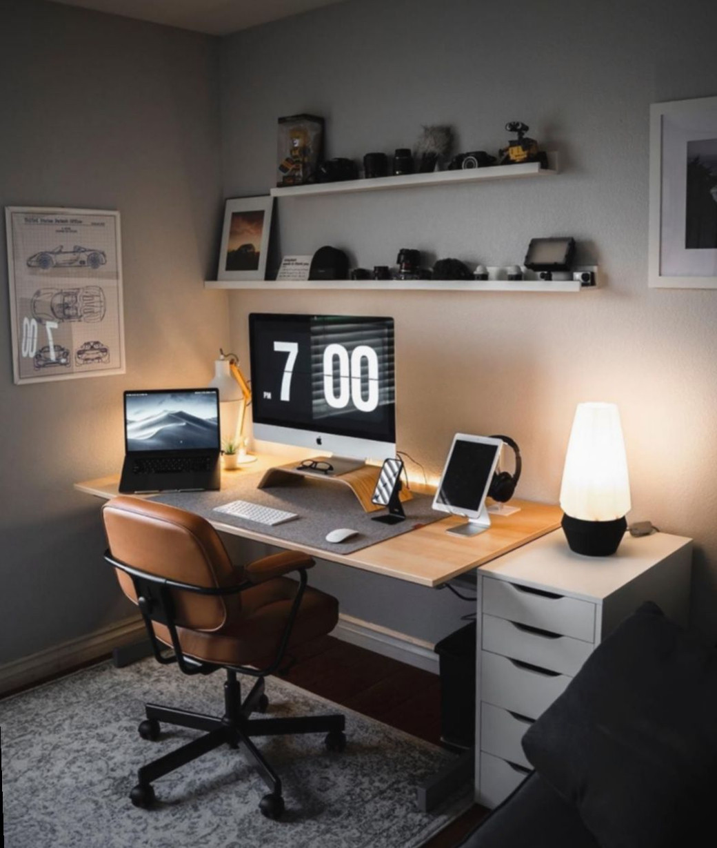 Tidy small home office with wooden desk, leather swivel chair, dual screens, shelves with cameras, and soft lighting.