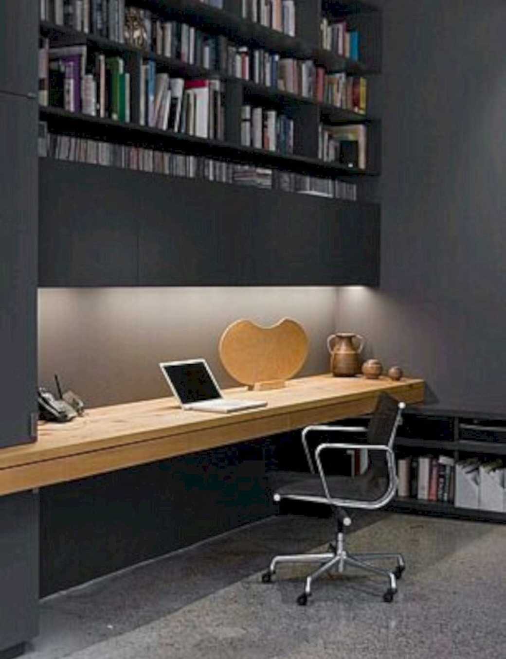 Compact home office with wooden floating desk, black swivel chair, shelves filled with books and decorative items.