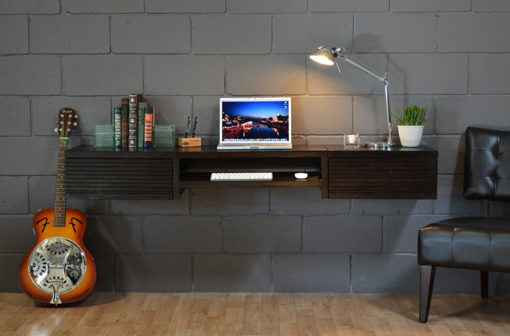 Floating dark wood desk mounted on grey brick wall with laptop, lamp, books, plant, guitar and black leather chair.