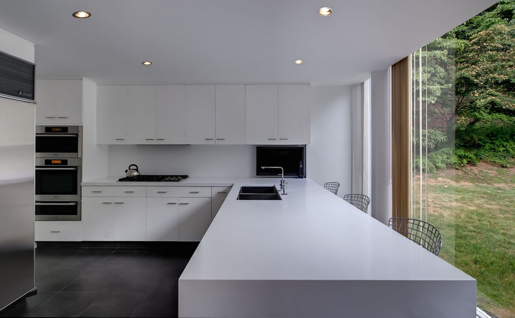 Modern white kitchen with sleek cabinetry, long island, built-in ovens, stainless steel appliances, and large window overlook