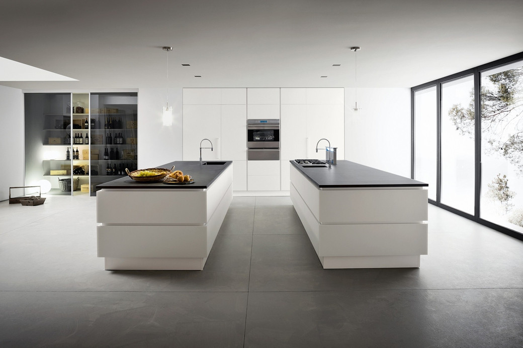 Minimalist white kitchen with two islands, black countertops, built-in oven, large windows, and a glass pantry.