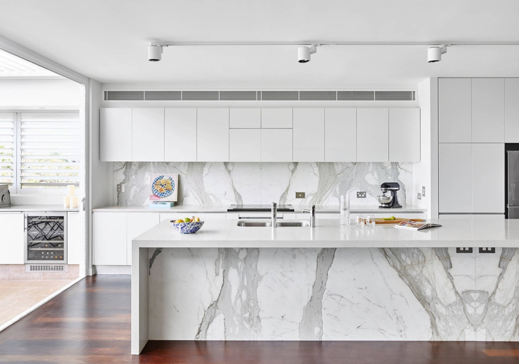 Bright modern white kitchen with marble countertops and backsplash, sleek cabinets, island with sink, and dark wooden floor.