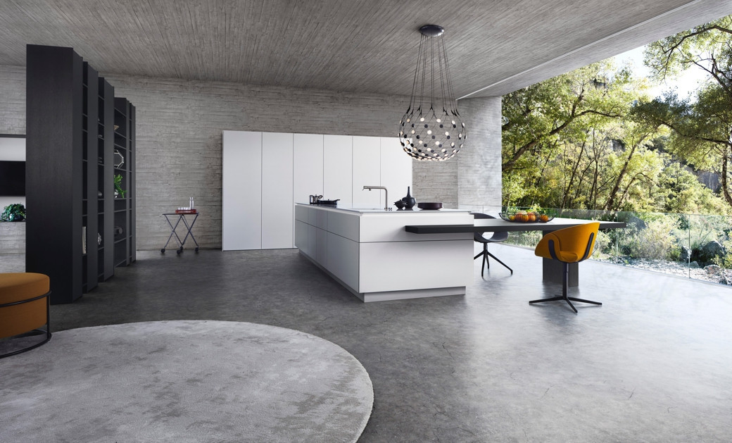 Modern white kitchen with central island, concrete floors and walls, large open window with forest view, and a yellow chair.