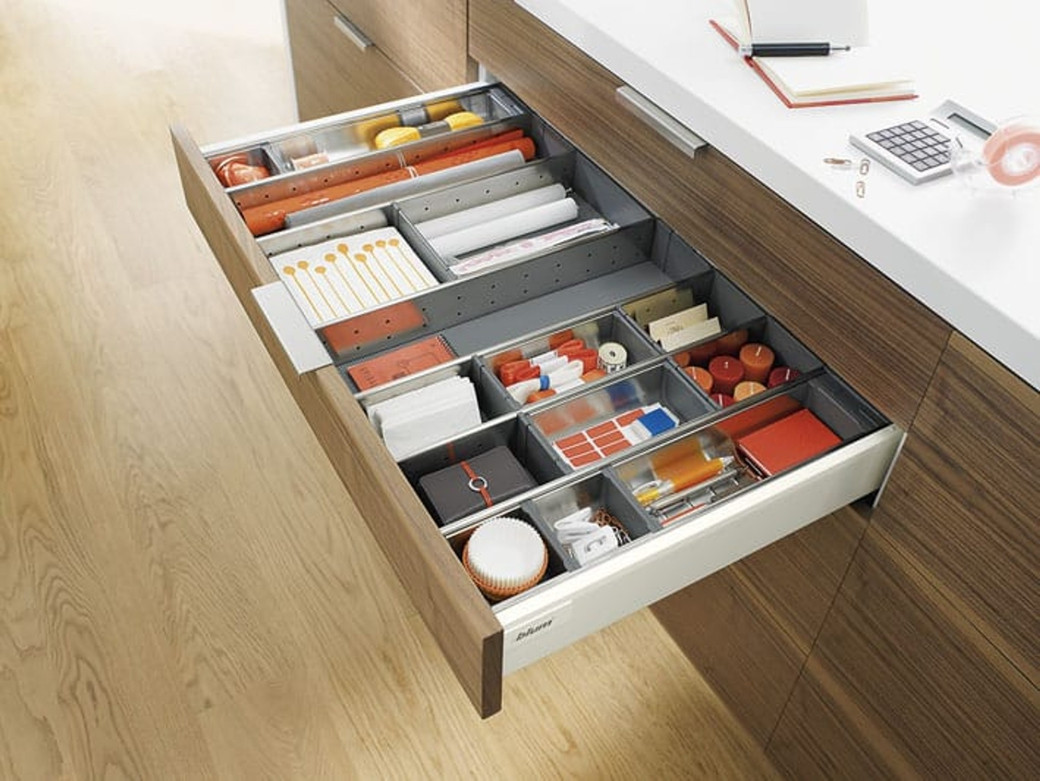 Open wooden kitchen drawer with multiple organised compartments holding candles, papers, and stationery items.