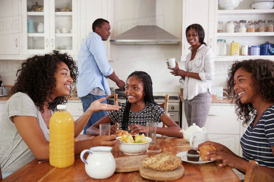 Family of five enjoying breakfast together in a kitchen with food and drinks on the wooden table.