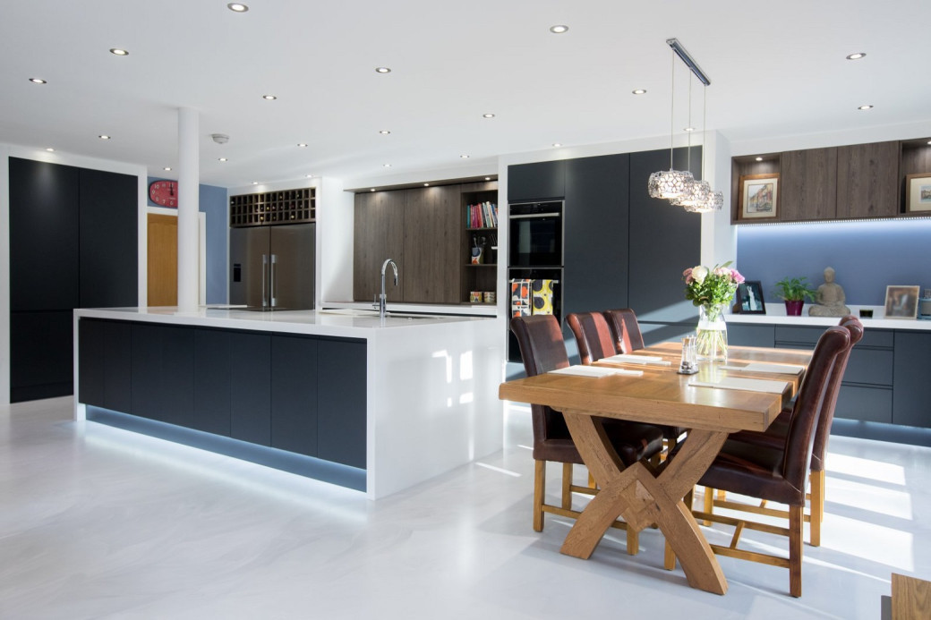 Modern kitchen with dark cabinets, white island with sink, wooden dining table, brown chairs, and pendant lighting.