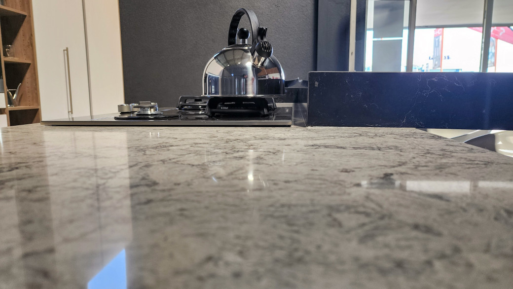 Close-up of a pale grey Caesarstone quartz kitchen countertop with a kettle and gas stove in background.
