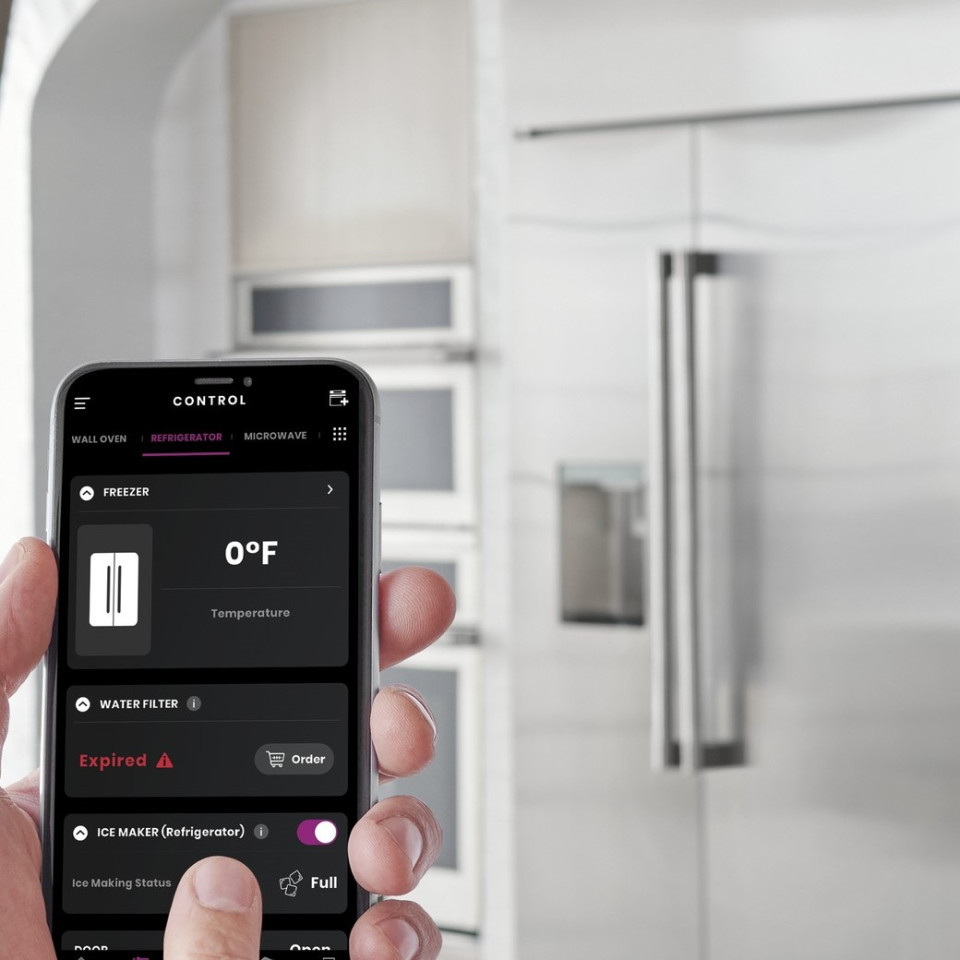 Hand holding smartphone displaying app controls for refrigerator temperature, water filter status, and ice maker in a modern