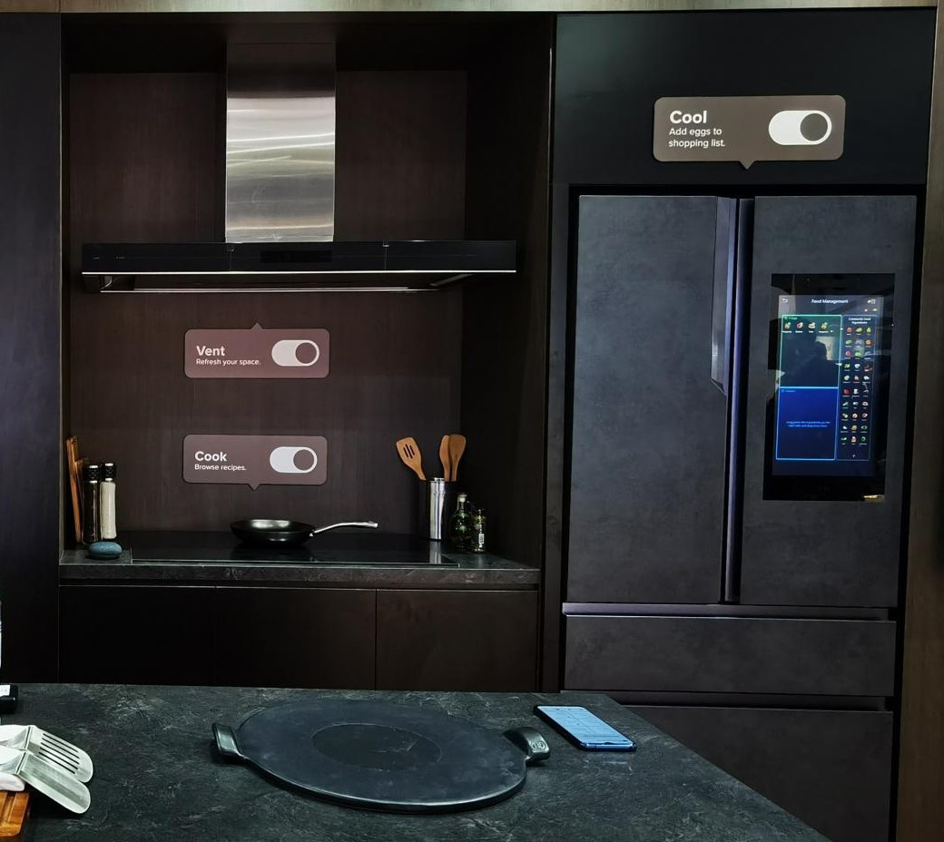Modern kitchen with dark cabinetry, a frying pan, cooking utensils, and a smart fridge displaying food management features.