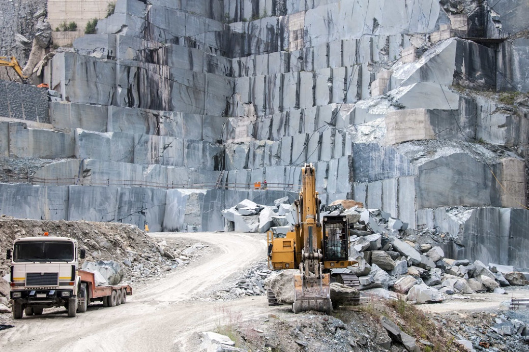 Open quarry with large cut stone blocks, a yellow excavator, and a white truck on a dirt road.