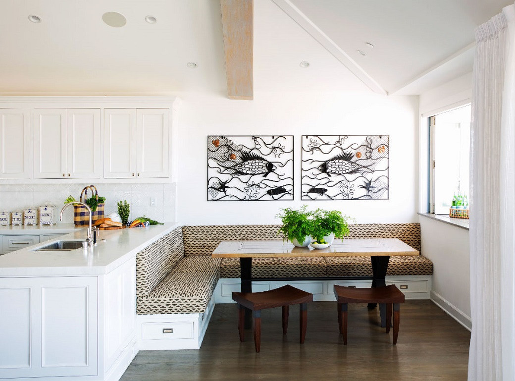 Bright kitchen corner with patterned L-shaped bench seating, wooden table, two stools, white cabinets, and fish art on wall.