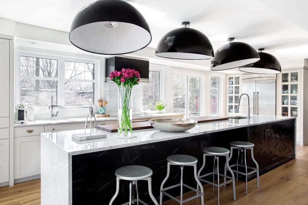 Modern kitchen with large marble island, four metal stools, black hanging pendant lights, and a vase of pink flowers.