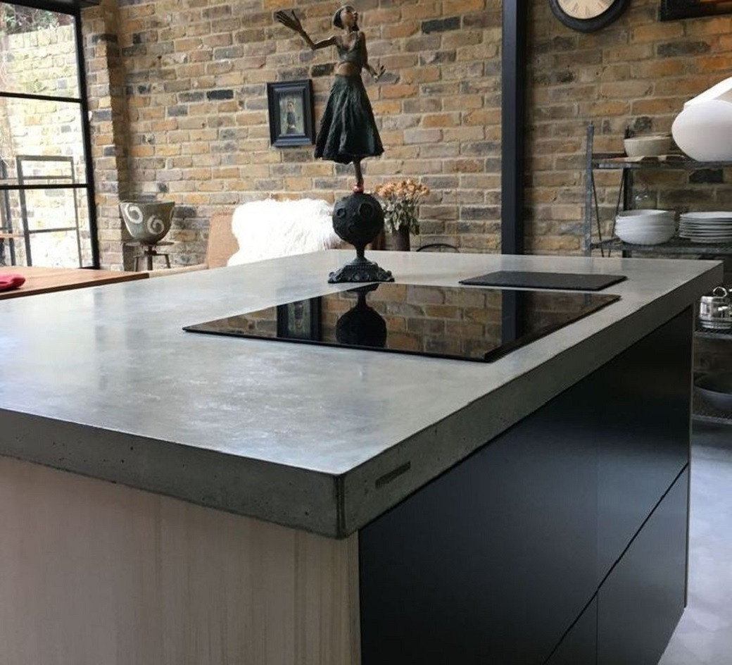 Concrete kitchen island countertop with built-in black induction stove, decorative figurine, and brick wall background.